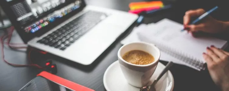 Un bureau avec un ordinateur, une tasse de café et une main qui écrit sur un carnet