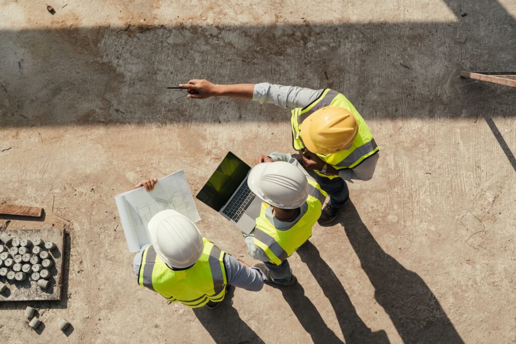 Trois hommes sont sur un chantier, ils tiennent des documents et un ordinateur